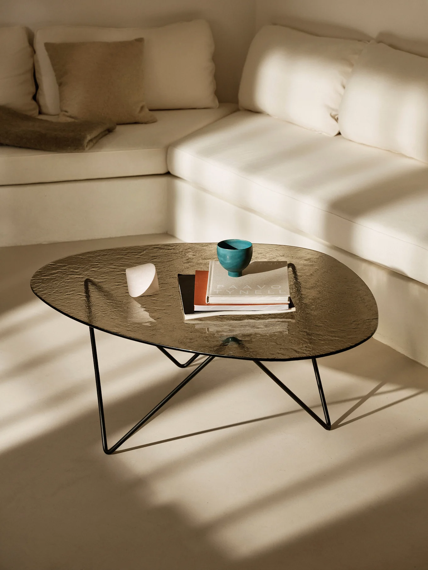 Dunkler Glastisch mit türkiser Schale, Büchern und weißem Objekt. Helles L-förmiges Sofa und Sonnenschatten.