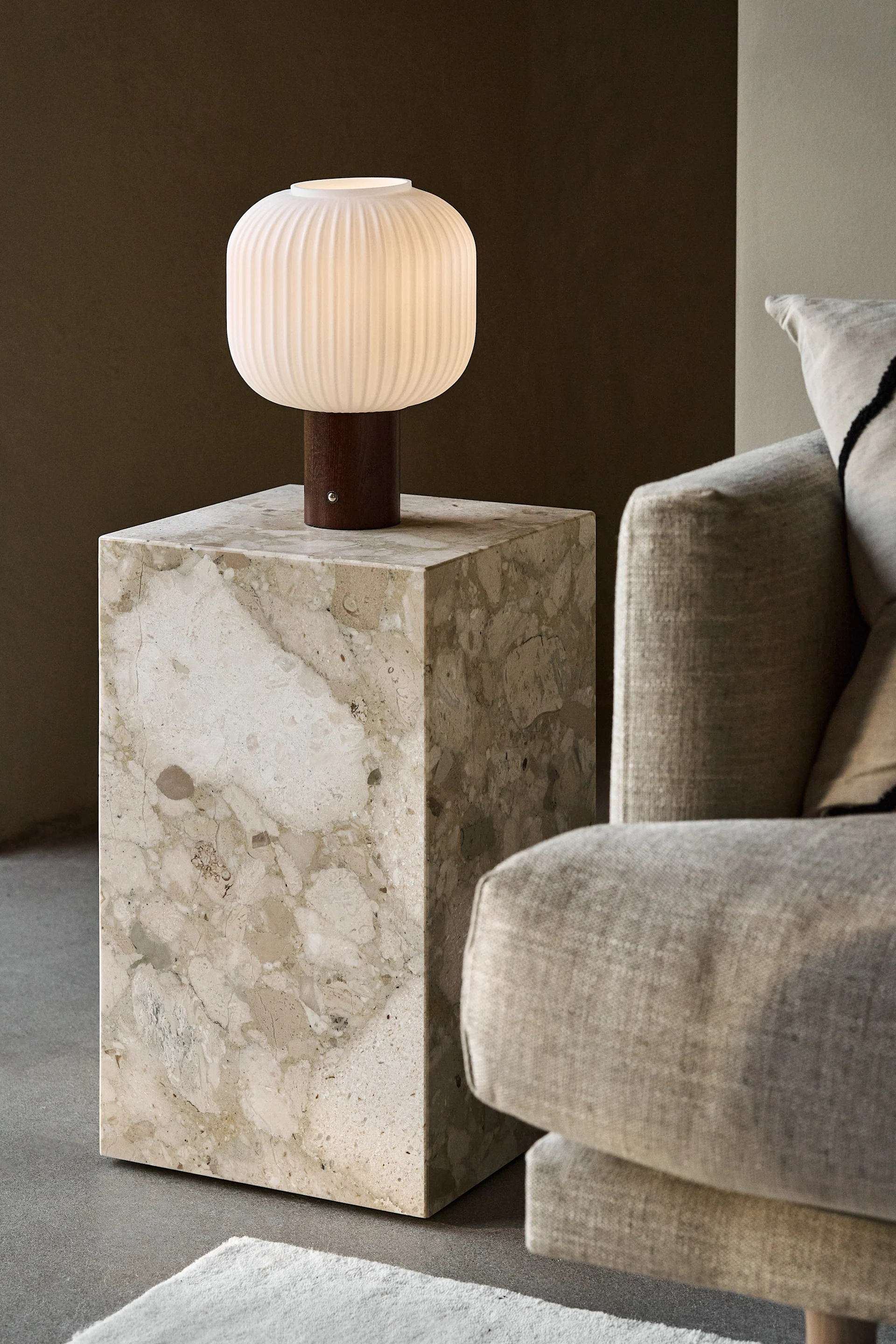 Eine beleuchtete, gerippte, weiße Tischlampe mit Holzsockel auf einem beigen Marmor-Beistelltisch neben einem hellen Sofa.