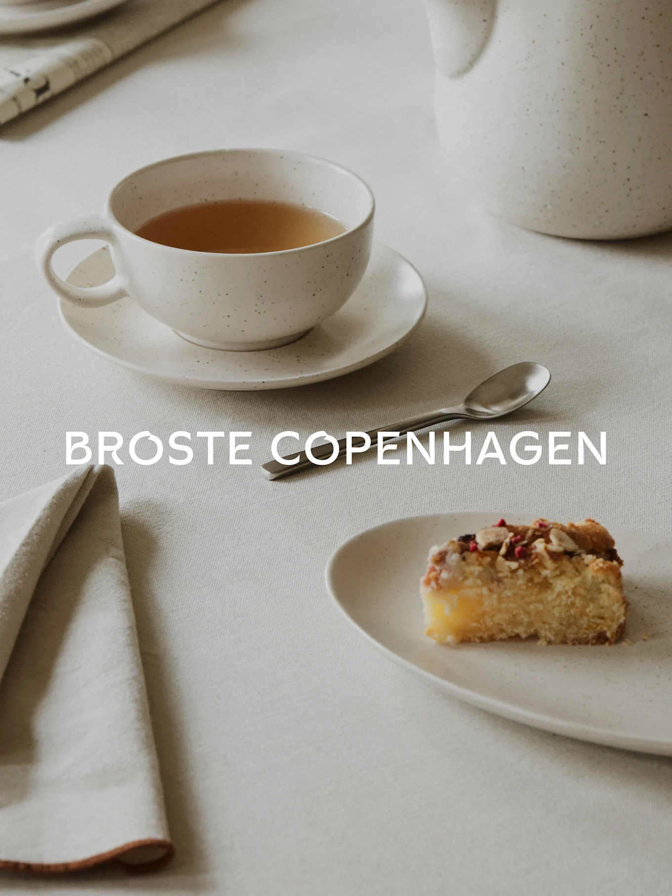 Tee in einer gesprenkelten weißen Tasse, ein Stück Kuchen mit Mandeln und Beeren auf einem passenden Teller, Löffel und Serviette auf Leinendecke. Text: BROSTE COPENHAGEN.