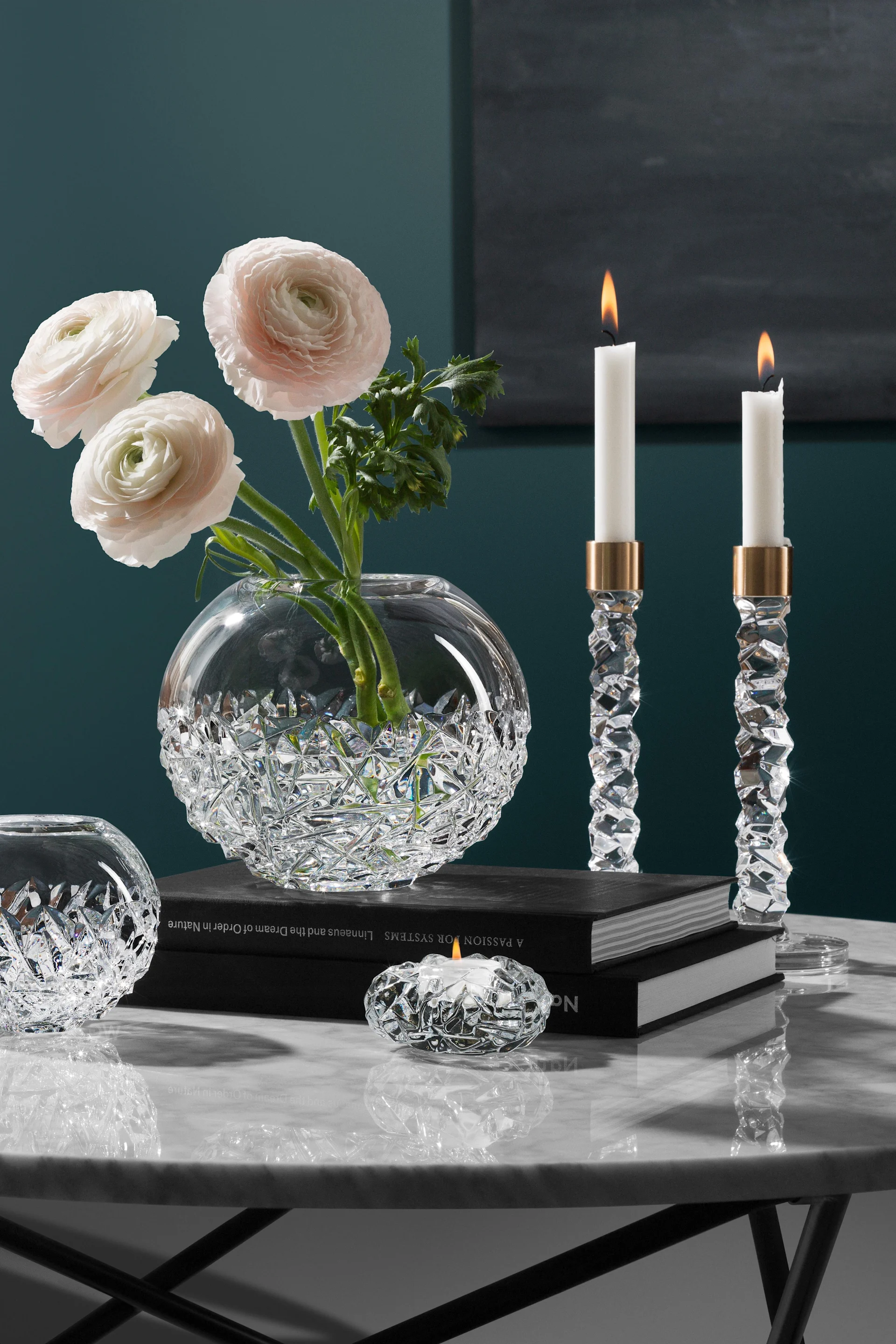 Elegante Kristallvase mit rosa Ranunkeln, zwei brennende Kristallleuchter und ein Teelichthalter auf einem Marmortisch.