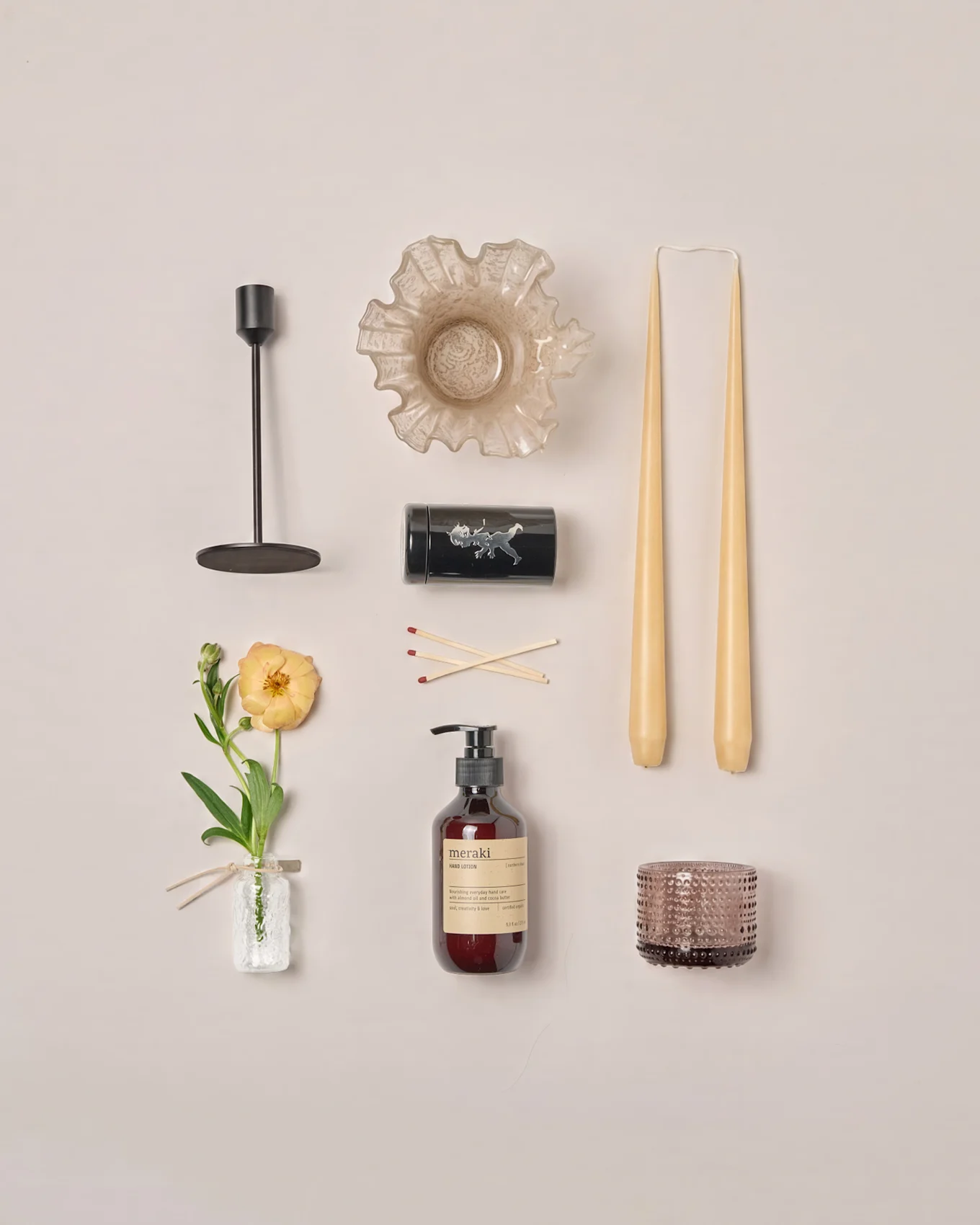 Flachansicht verschiedener Wohnaccessoires: Kerzenleuchter, Rüschenschale, konische Kerzen, Streichhölzer, Lotion, Teelichthalter und eine Ranunkelblume.