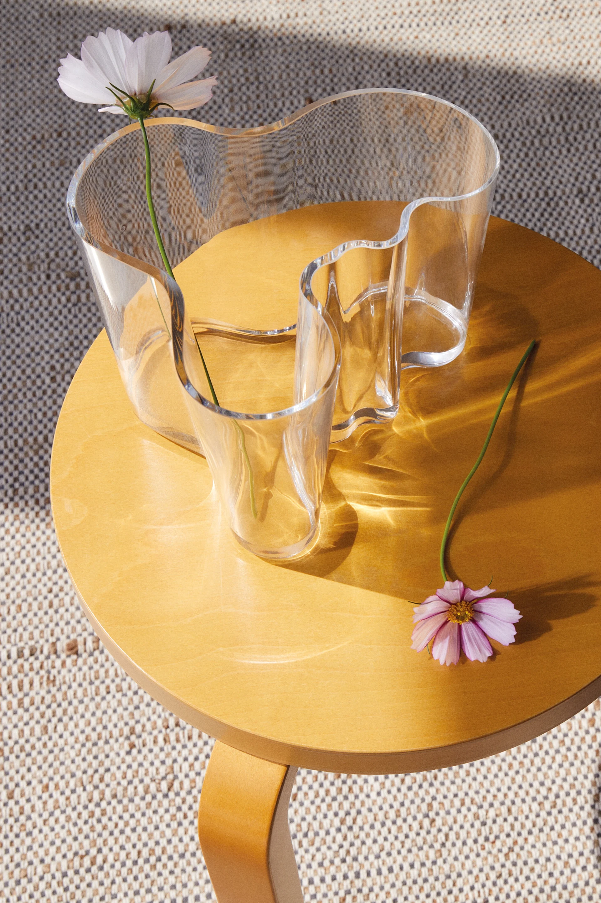 Eine klare, wellenförmige Glasvase mit weißer Blume auf einem runden Holztisch, daneben eine rosa Blume. Sonnenlicht wirft Muster.