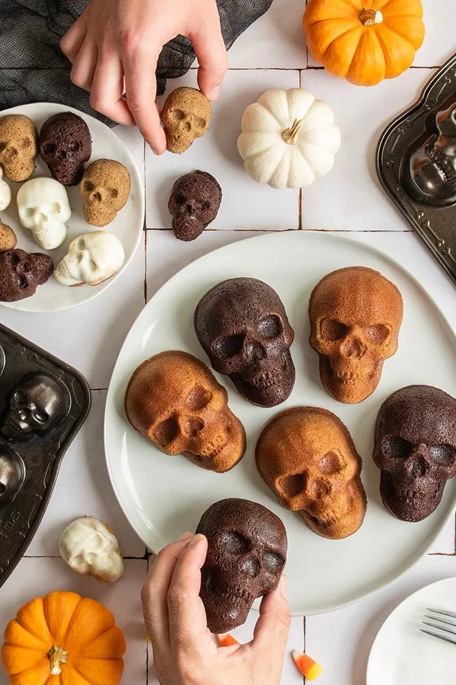 Hände arrangieren verschiedene totenkopf-förmige Halloween-Kuchen (Dunkel-Schokolade, Kürbis-Gewürz, weiße Schokolade) mit Mini-Kürbissen und Candy Corn.