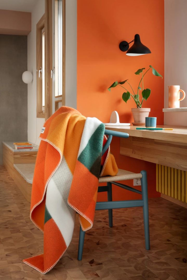 Farbtrends 2024: Zu den Trendfarben 2024 gehört auch Orange. Hier sehen Sie die gemusterte Mikkel-Decke von Røros Tweed in Orange, Grün und Weiß über einem Stuhl an einem Schreibtisch.