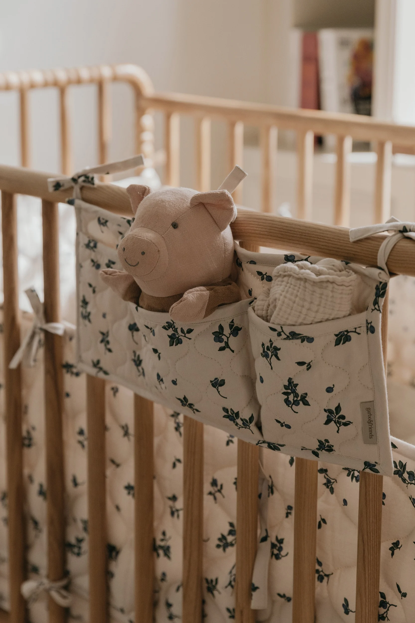 Ein rosa Plüschschwein und ein weißes Musselintuch in einem creme-blauen Blumen-Organizer, der an einem Holzbett hängt.