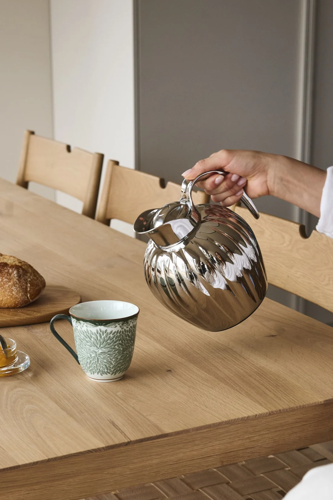 Eine Hand gießt aus einer silbernen geriffelten Thermoskanne in eine grüne Tasse auf einem Holztisch mit Brot und Marmelade.