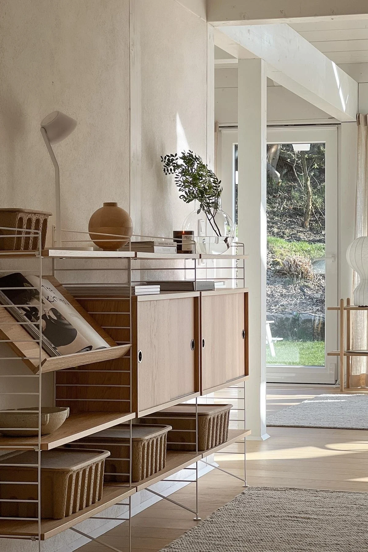 Helles, modernes Zimmer mit Regalsystem aus hellem Holz und weißen Rahmen, gefüllt mit Vase, Büchern, Pflanzen und Körben, mit Gartenblick.