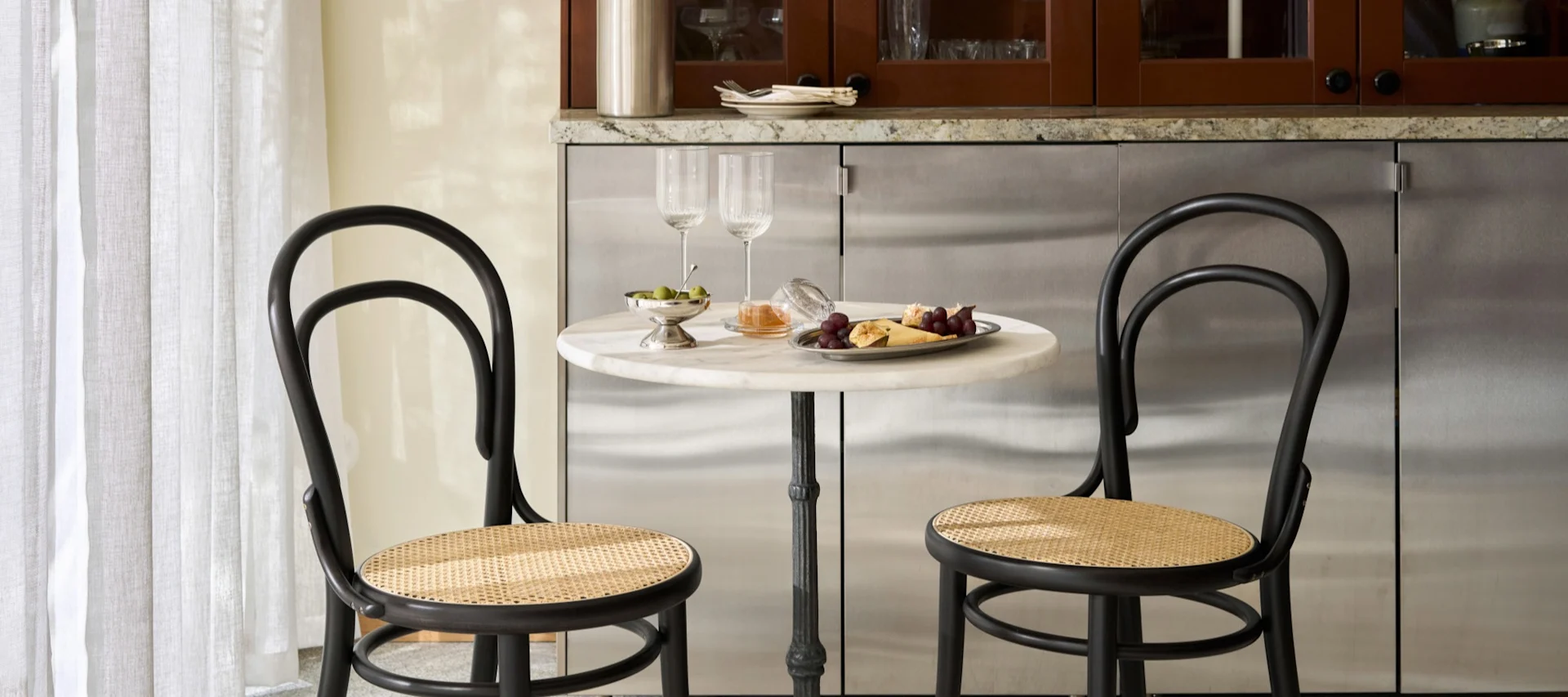 Zwei schwarze Bistrostühle mit Rattansitzen an einem runden Marmortisch mit Weingläsern, Oliven, Trauben und Käse.