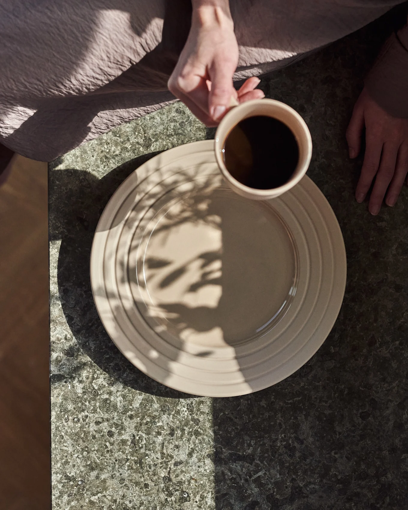 Draufsicht: Hände, eine hält eine beige Tasse mit schwarzem Kaffee über einem beigen Teller auf dunklem Steintisch, mit Blattschatten.