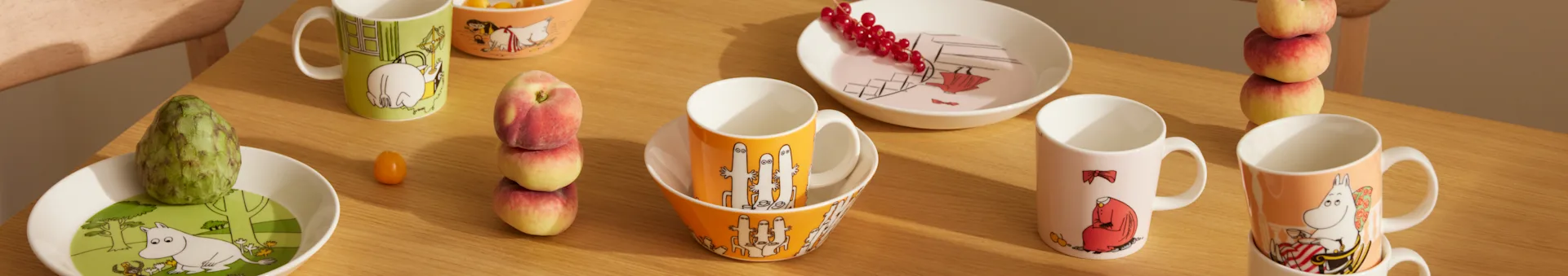 Moomin-Geschirr in verschiedenen Farben und Designs, mit Tassen, Schüsseln und Tellern, dazu frisches Obst auf einem Holztisch.