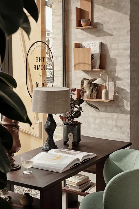 Ein gemütlicher Raum mit einem dunklen Holztisch, einem offenen Kunstbuch, einem Glas Wein, einer grauen Lampe und einer Pflanze. Holzregale mit Dekorationsgegenständen sind an der Wand.