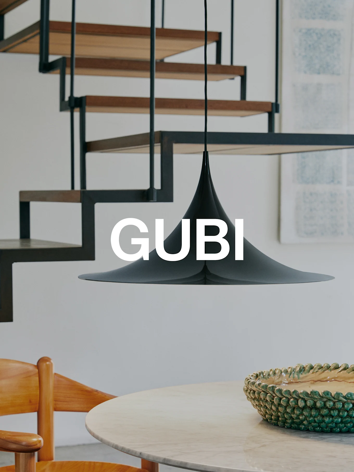 Eine schwarze konische Pendelleuchte über einem Marmortisch mit einer grünen Schale und einem Holzstuhl. Eine moderne Treppe im Hintergrund. Text 'GUBI'.