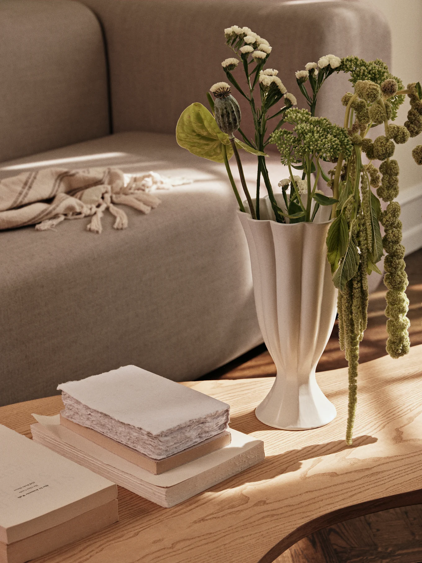 Eine weiße geriffelte Vase mit grünen Pflanzen und ein Stapel Bücher auf einem Holztisch, mit einem Sofa im Hintergrund.