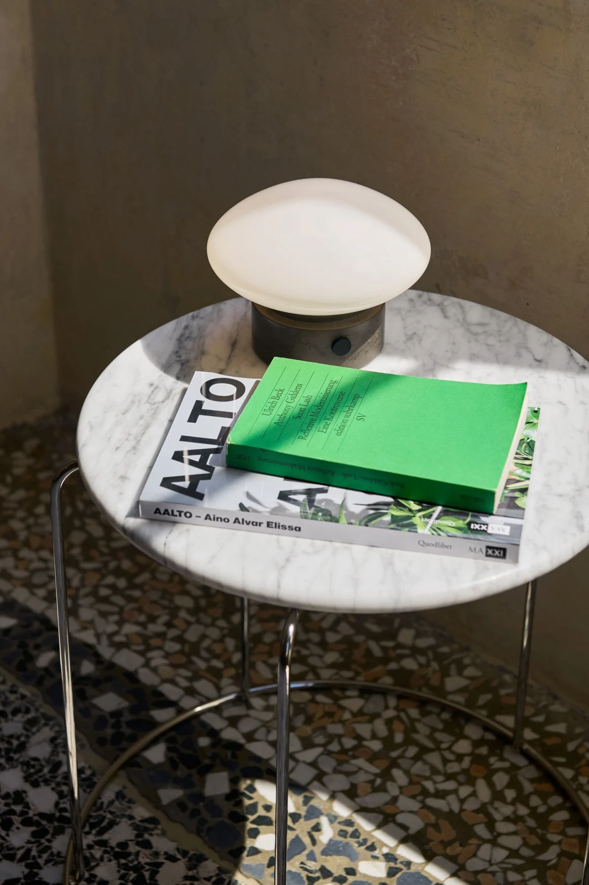 Die Akku-Tischleuchte Mist AP12 von &tradition steht zusammen mit Büchern auf einem kleinen runden Marmortisch. 