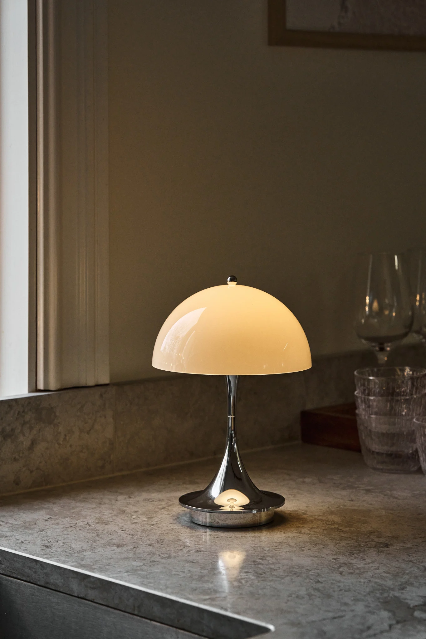 Tischlampe aus poliertem Chrom mit leuchtendem cremefarbenem Kuppelschirm auf einer gesprenkelten grauen Arbeitsplatte, neben einem Fenster und verschwommenen Gläsern.