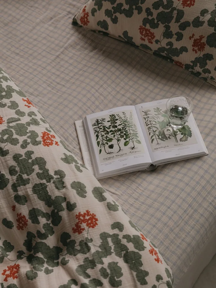 Ein offenes Botanikbuch und ein Glas Wasser auf einem Bett mit karierter Bettdecke und floral gemusterten Kissen.