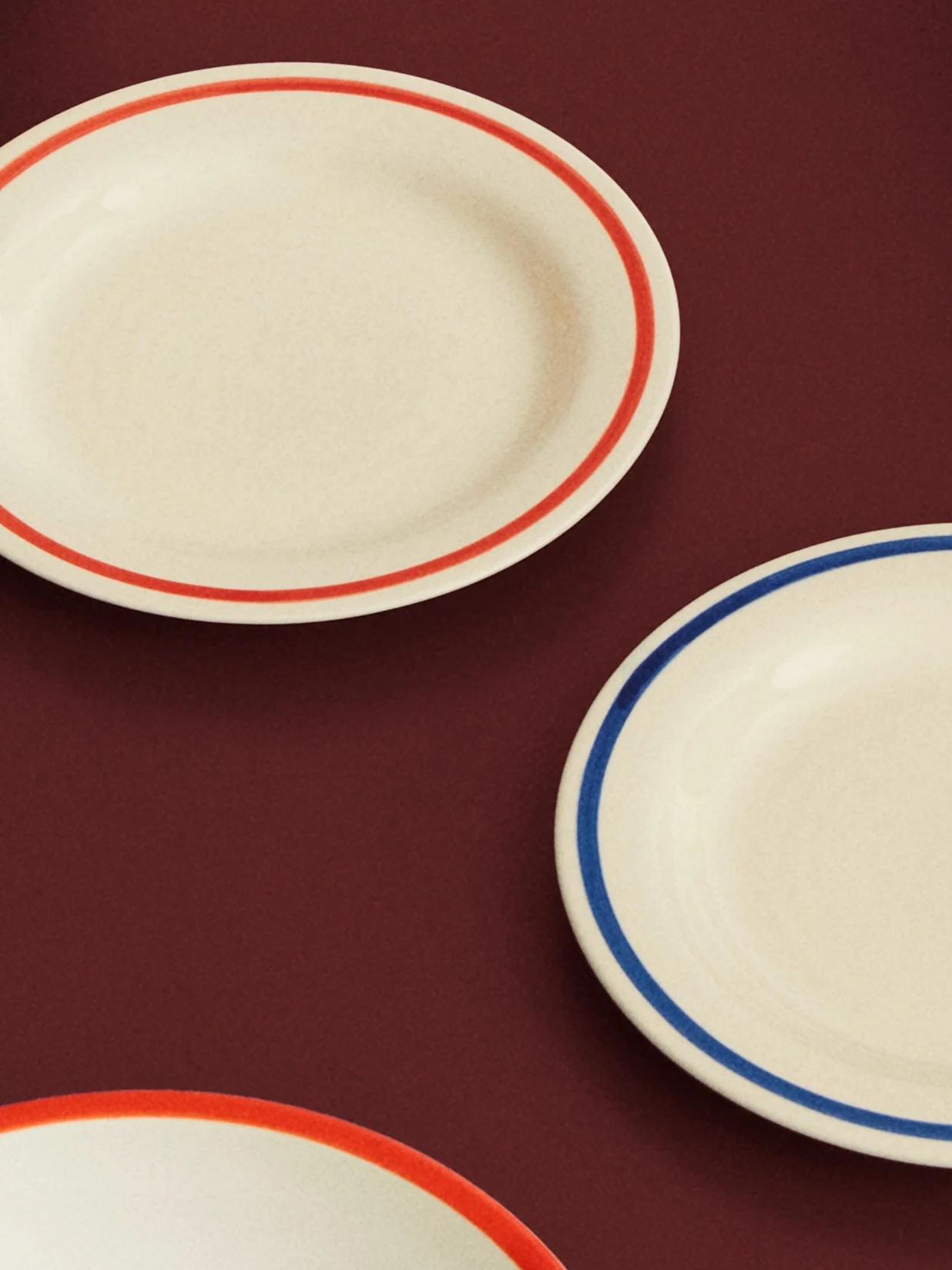 Drei cremefarbene ovale Teller mit roten und blauen Rändern auf einer dunkelroten Oberfläche.