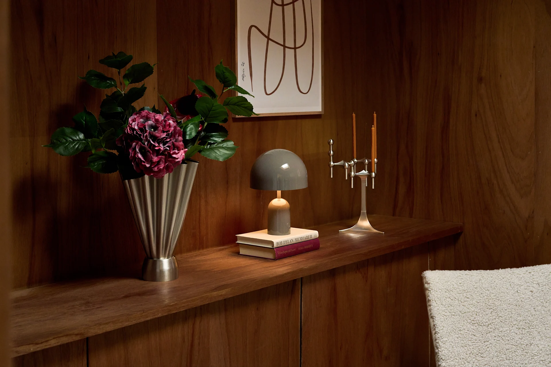 Ein Holzregal zeigt eine silberne Vase mit lila Blumen, eine graue Pilzlampe auf Büchern, und einen silbernen Kerzenständer mit zwei brennenden Kerzen.