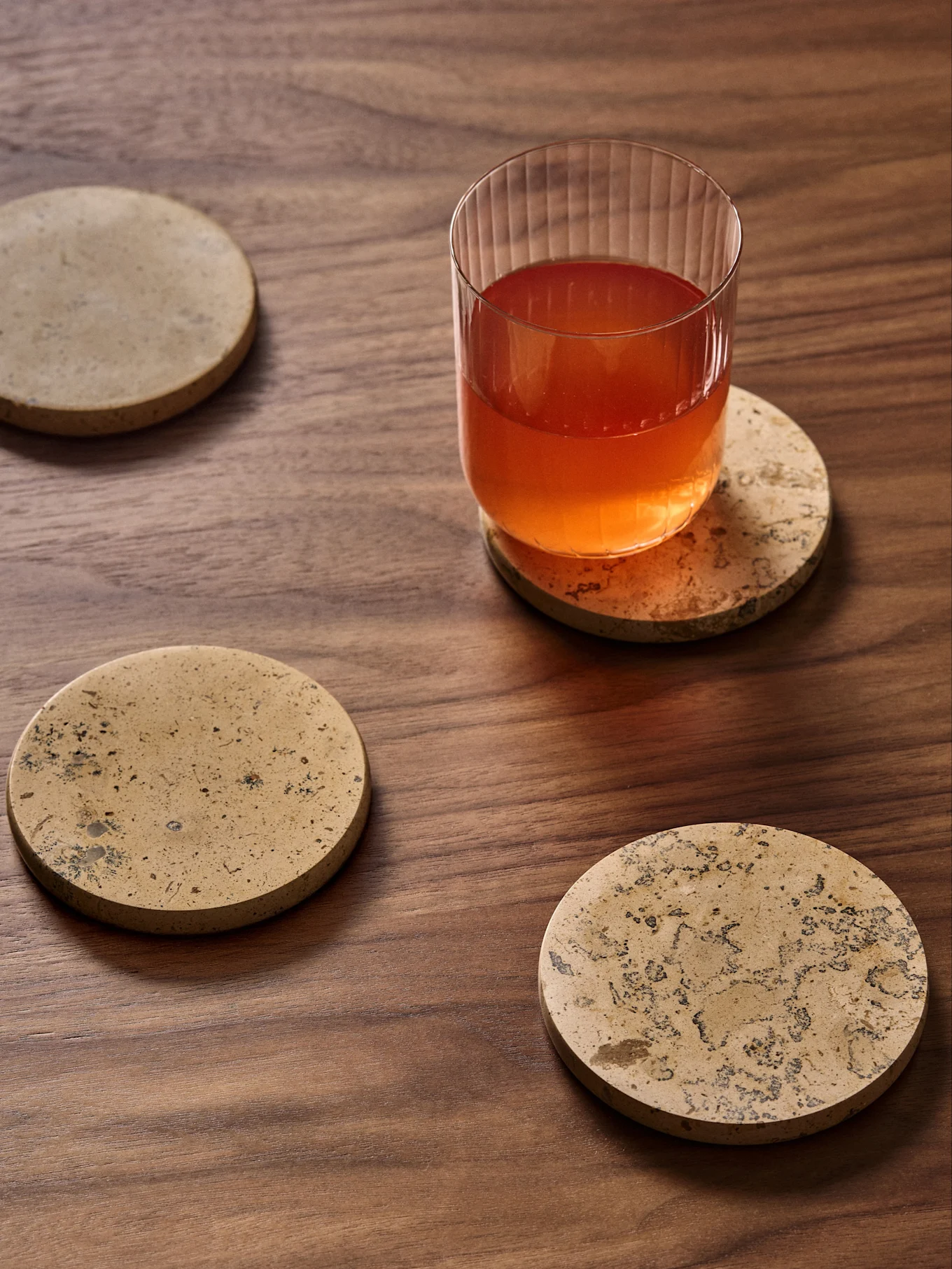 Ein Glas mit rötlichem Getränk auf einem runden Marmoruntersetzer, mit drei weiteren ähnlichen Untersetzern auf einem dunklen Holztisch verteilt.