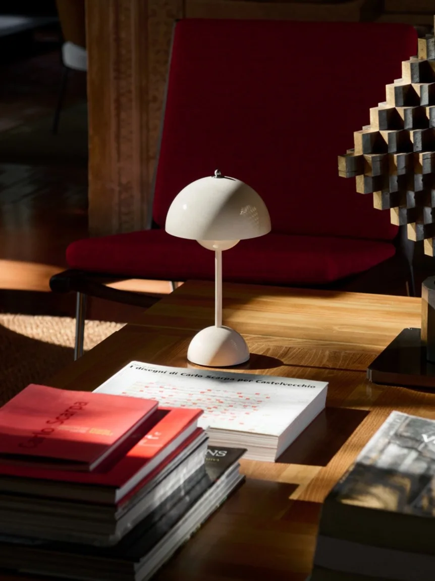 Eine weiße Pilz-Tischlampe, Bücher und eine geometrische Skulptur auf einem Holztisch, mit einem roten Sessel im Hintergrund.