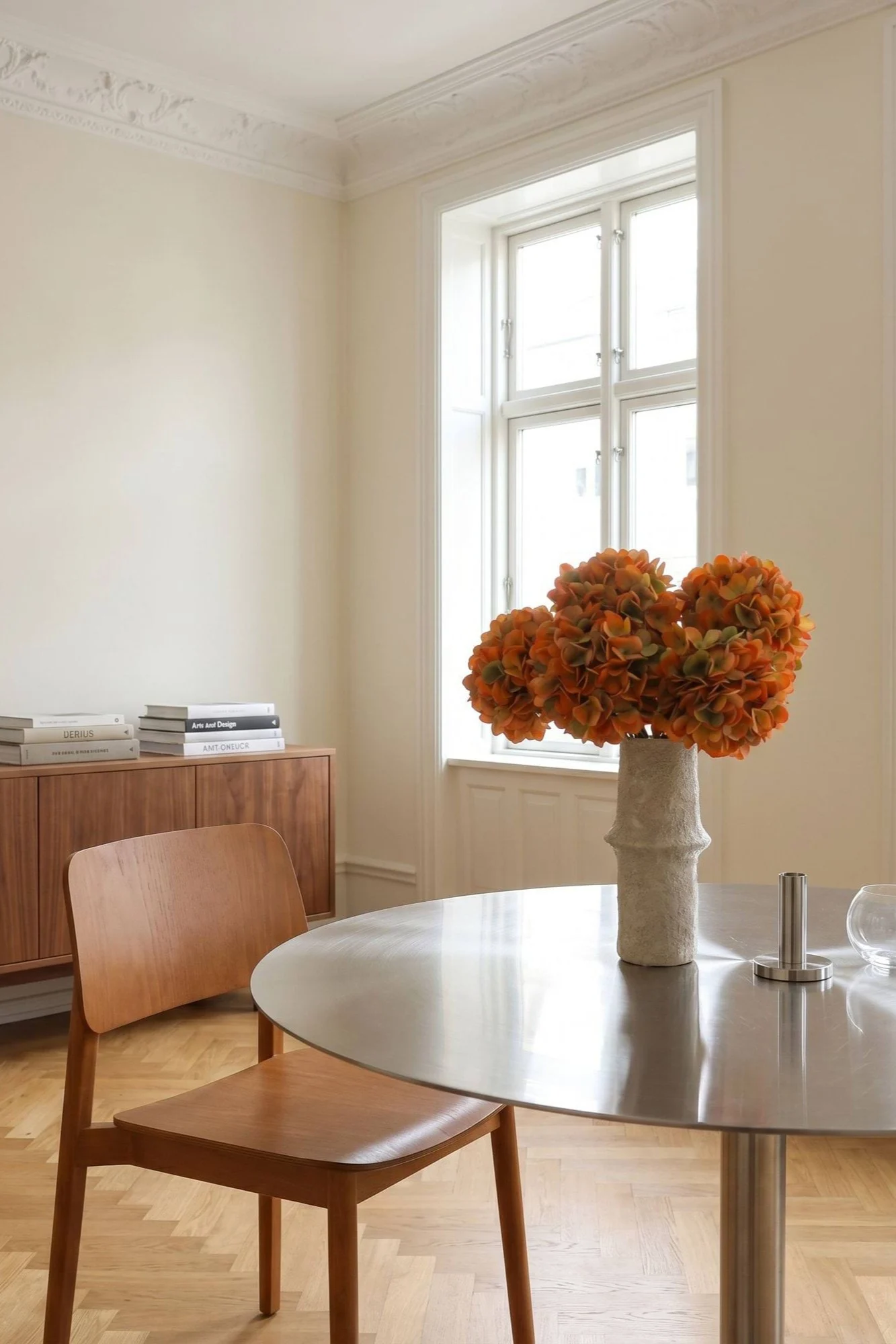 Ein helles Esszimmer mit einem runden Metalltisch, einem Holzstuhl und einer Vase mit orangefarbenen Blumen.