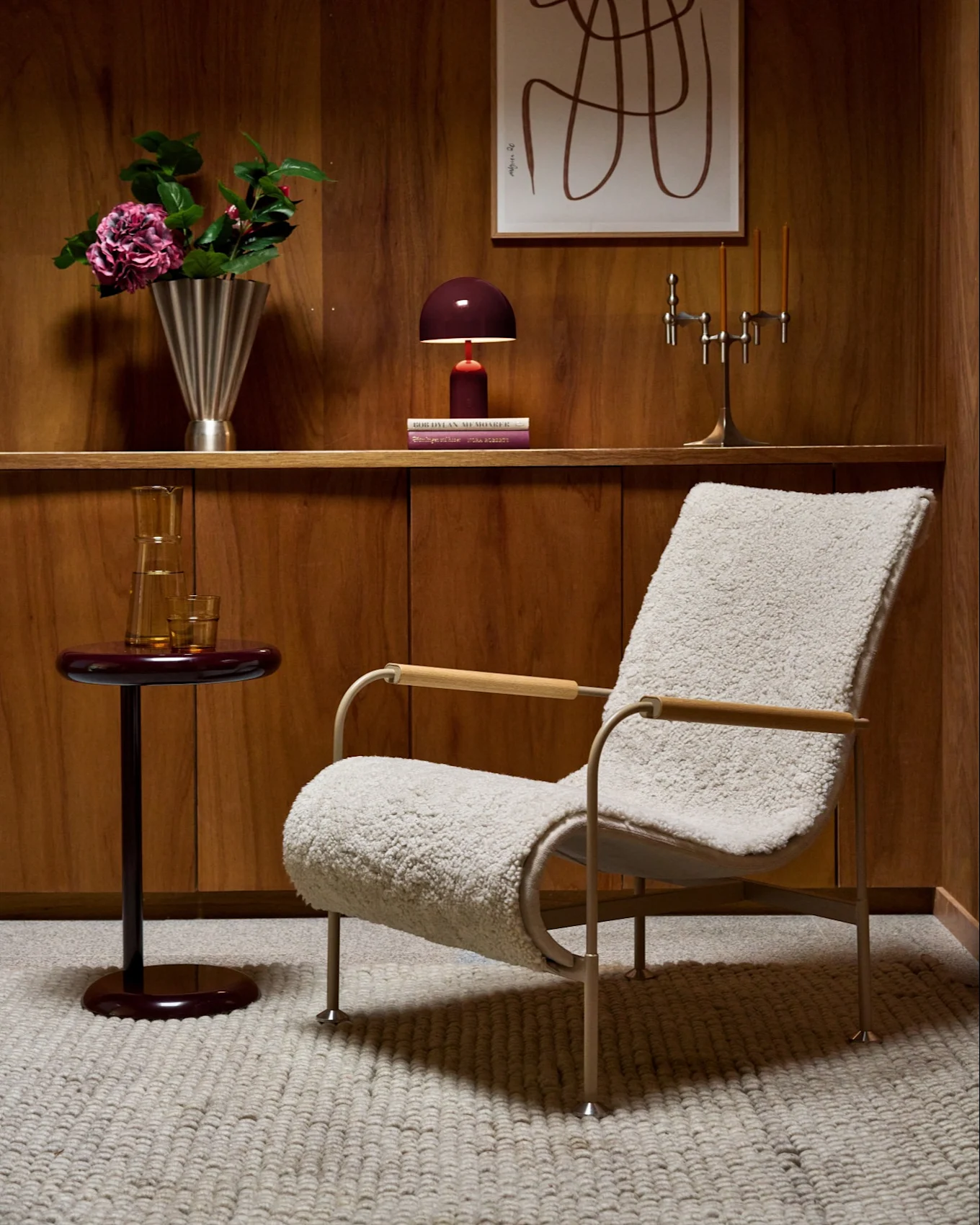 Holzgetäfeltes Zimmer mit flauschigem Sessel, rotem Beistelltisch, Regal mit Vase, Lampe, Kerzenständer und abstrakter Kunst.