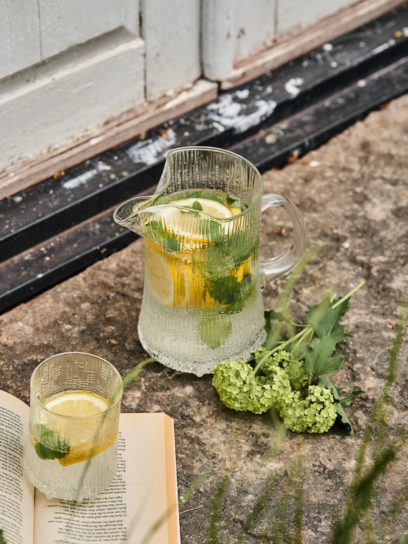 Gerippte Glaskanne und passendes Glas mit Zitronen- und Minzwasser, neben einem offenen Buch und grünen Blumen auf Stein.