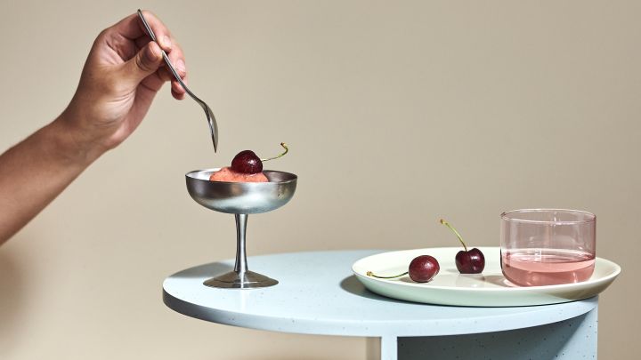 Die Wohntrends 2022 umfassen sanfte Pastellfarben, inspiriert von den 80er Jahren, hier ruht das Tint Trinkglas und die Italian Ice Cup Dessertschale, beide von HAY, auf dem Teema-Teller von Iittala, begleitet von Eis und Kirschen.