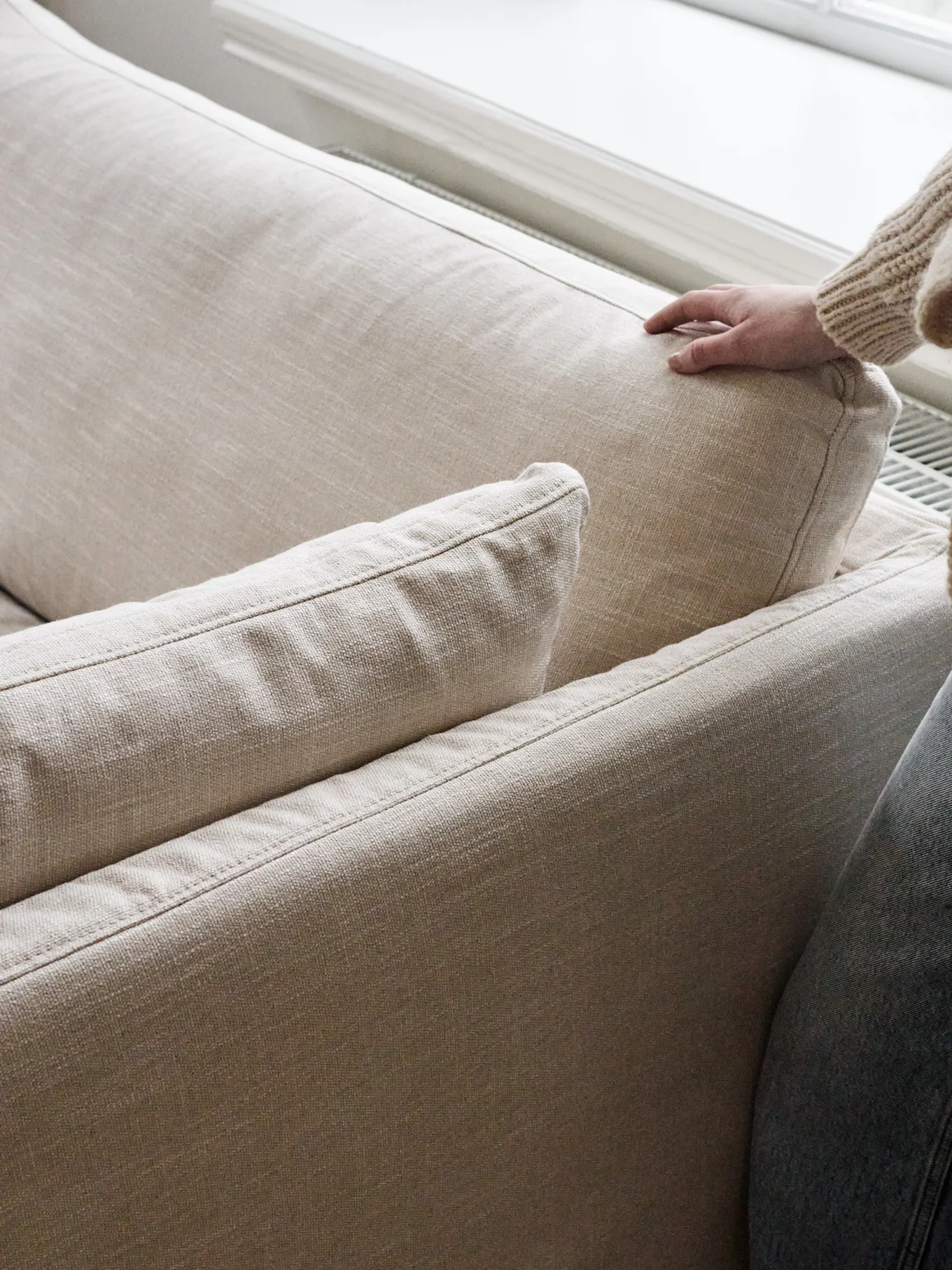 Eine Hand in einem grob gestrickten Pullover drückt sanft in das Rückenkissen eines hellbeigen Sofas.