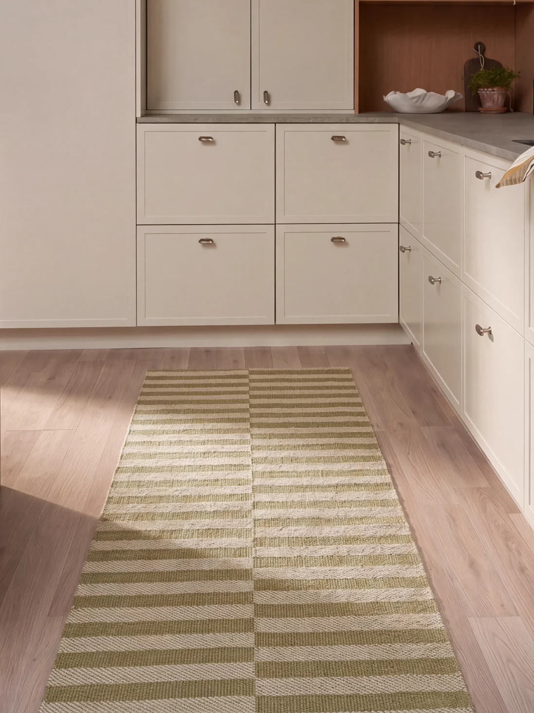 Eine Küche mit hellen Schränken, Holzboden und einem langen rechteckigen Teppich mit grünen und weißen Querstreifen.