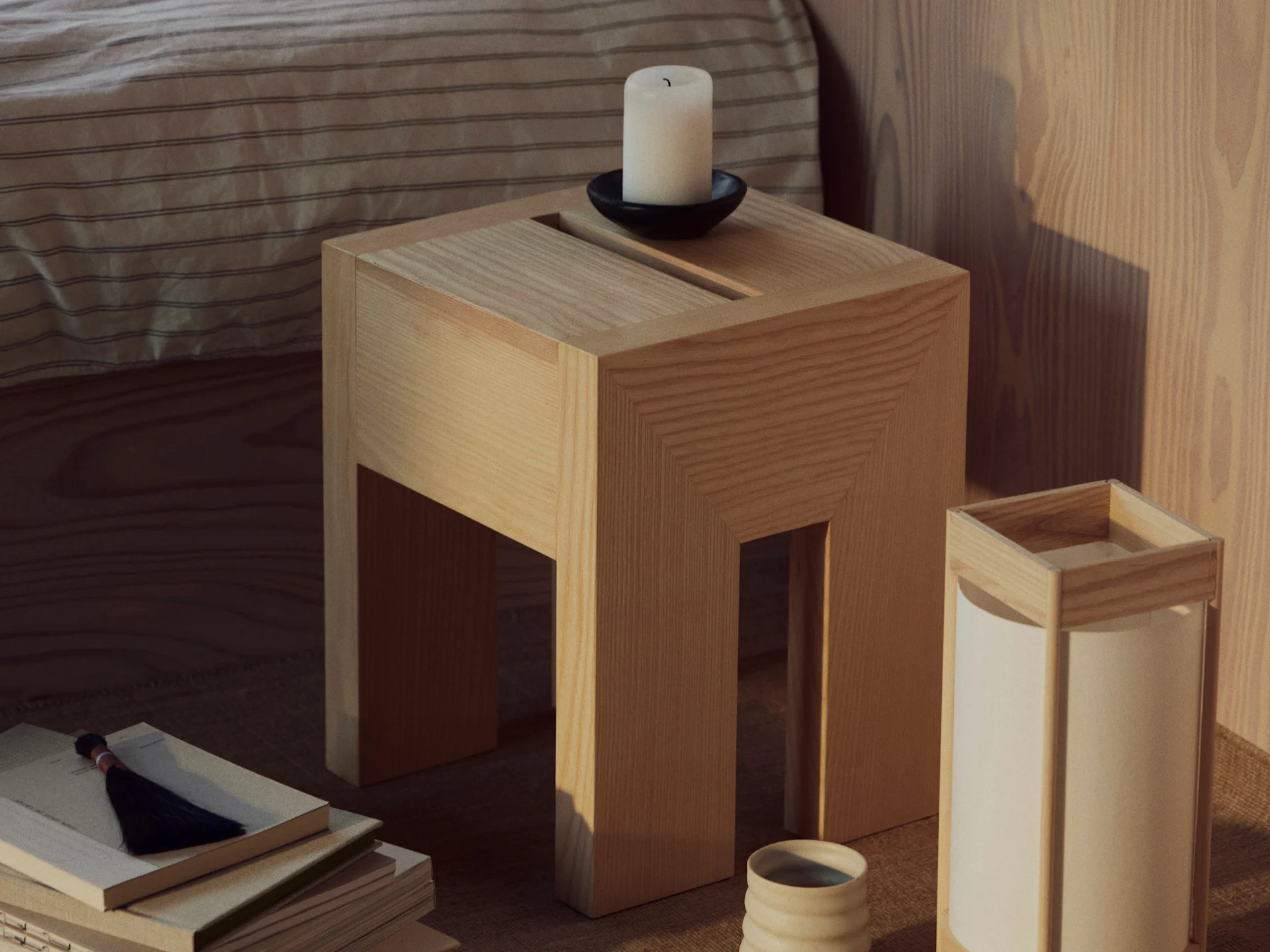 Ein Holznebentisch mit einer weißen Kerze in einem schwarzen Halter, einem Stapel Bücher mit Quaste und einer Lampe mit Keramikbecher.