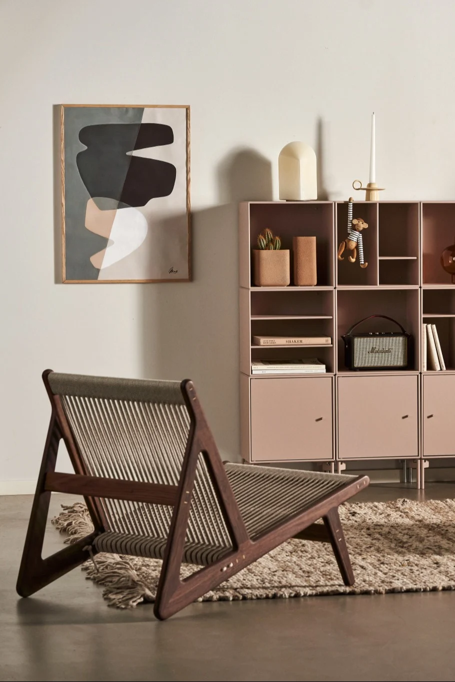 Ein modernes Interieur mit einem Lounge-Stuhl aus Holz mit Seilgeflecht, einem pinken Modulschrank und abstrakter Wandkunst.