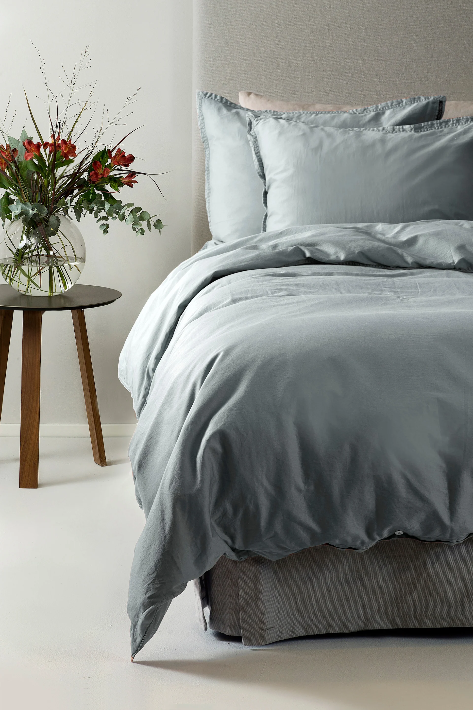 Ein Bett mit hellblauer Bettwäsche und zwei Kissen, daneben ein Holztisch mit einer Vase roter Blumen.