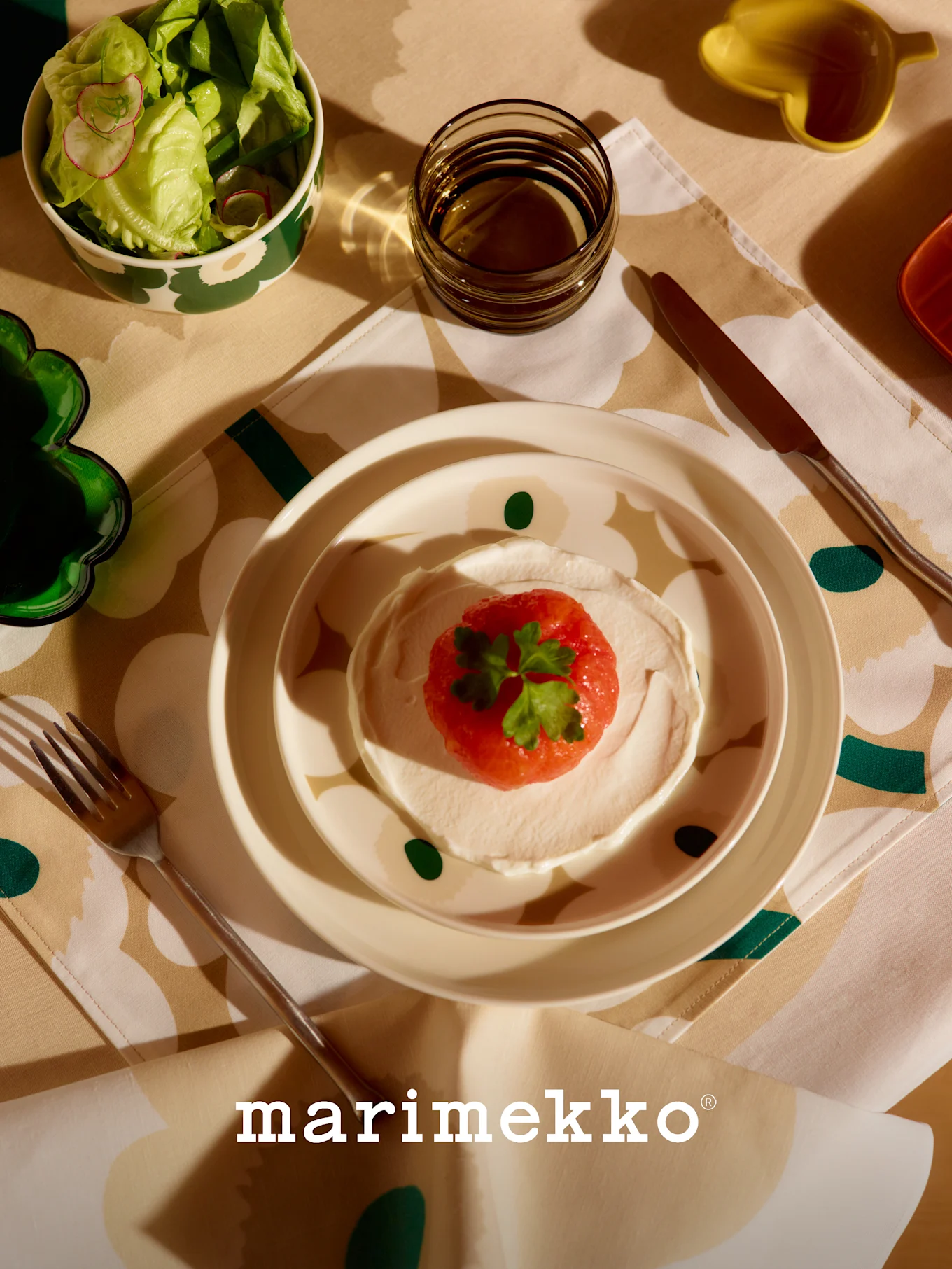 Marimekko Tischgedeck mit gemusterter Salatschüssel, Gericht mit roter Frucht und Creme, Glas und Besteck auf einer floralen Tischdecke.