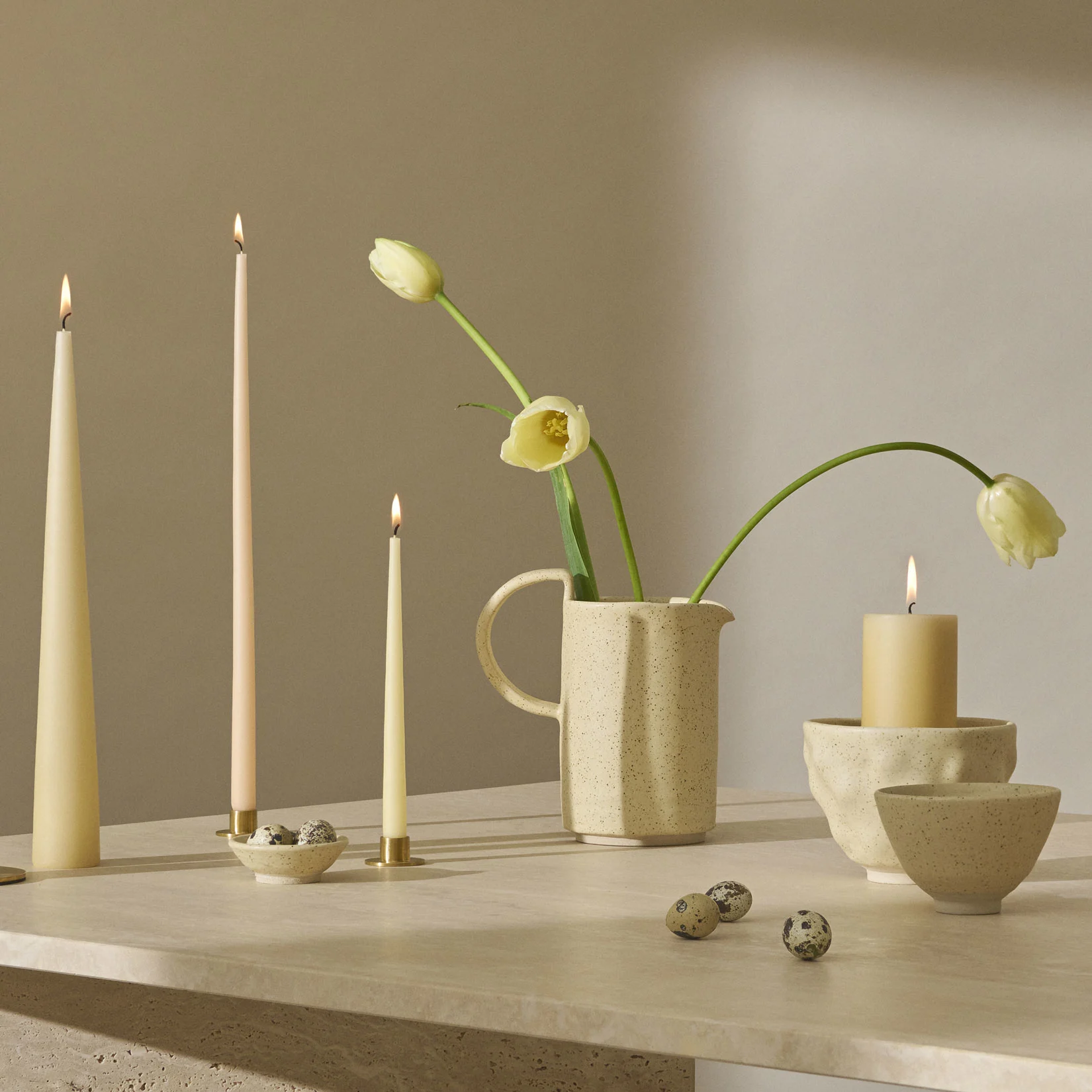 Ein Stillleben mit brennenden Kerzen, hellgelben Tulpen in einem gesprenkelten Krug und Wachteleiern auf einer strukturierten Oberfläche.