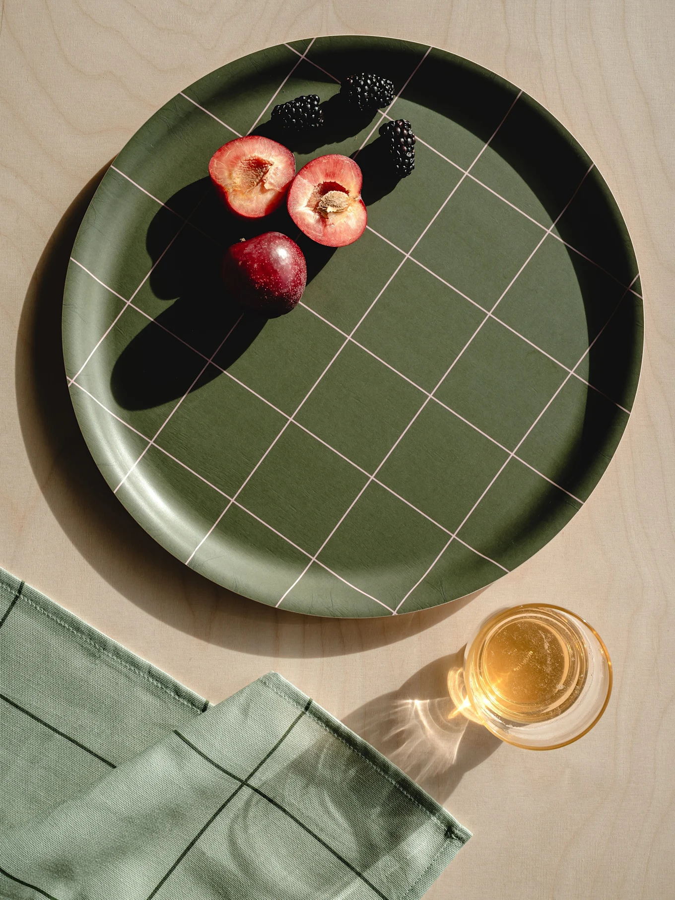 Dunkelgrünes kariertes Tablett mit roten Pflaumen, Brombeeren, hellgrüner Serviette und einem Glas Saft auf Holz.