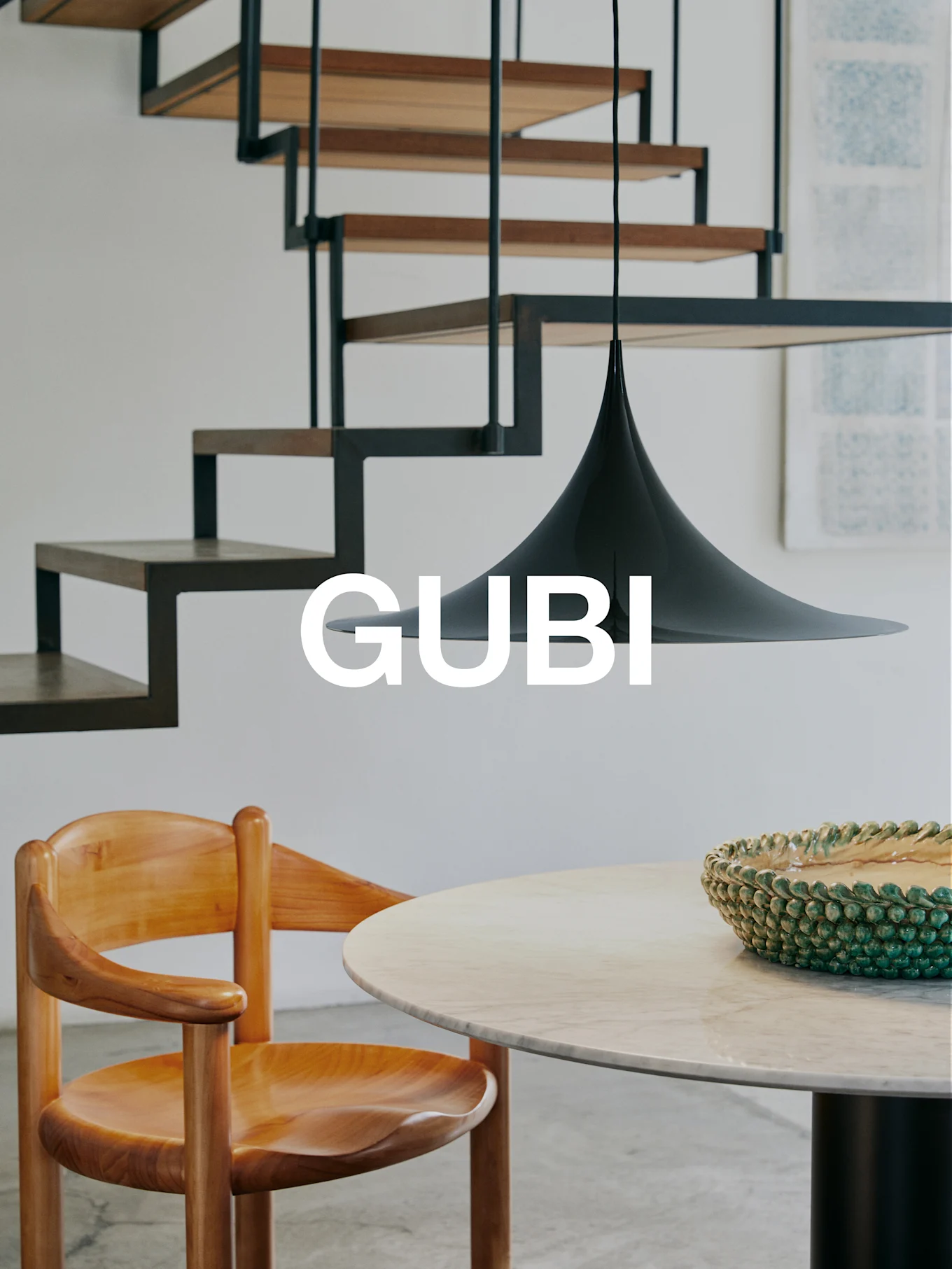 GUBI-Logo in einem modernen Interieur mit Holzstuhl, Marmortisch, grüner Schale, schwarzer Pendelleuchte und moderner Treppe.