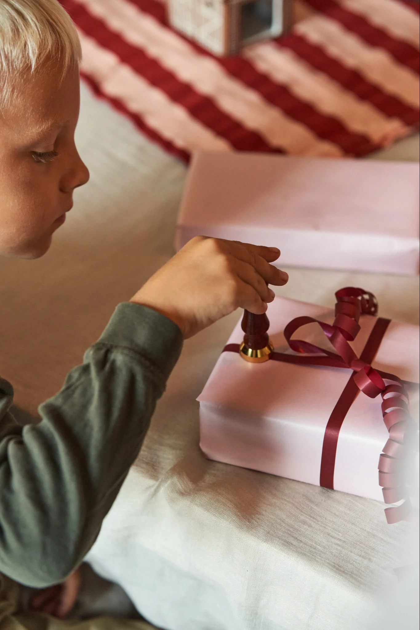 Es ist der erste Advent, und ein Junge stempelt ein Weihnachtsgeschenk, das in rosa Papier und rote Geschenkband eingepackt ist.