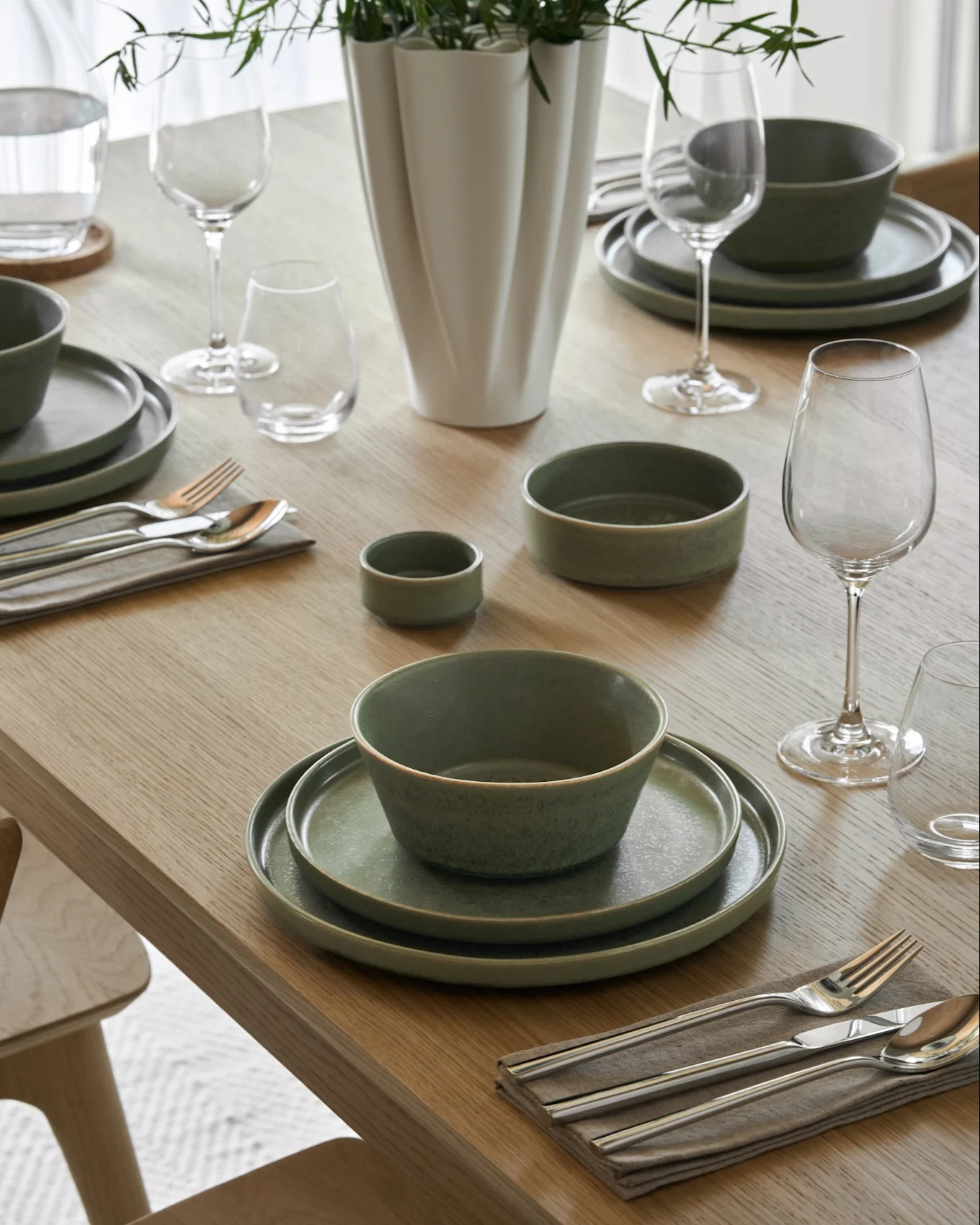 Esstisch gedeckt mit cremigen Suppenschalen, passenden Tellern, Wein- und Wassergläsern, Besteck und einer Vase mit grünem Laub.