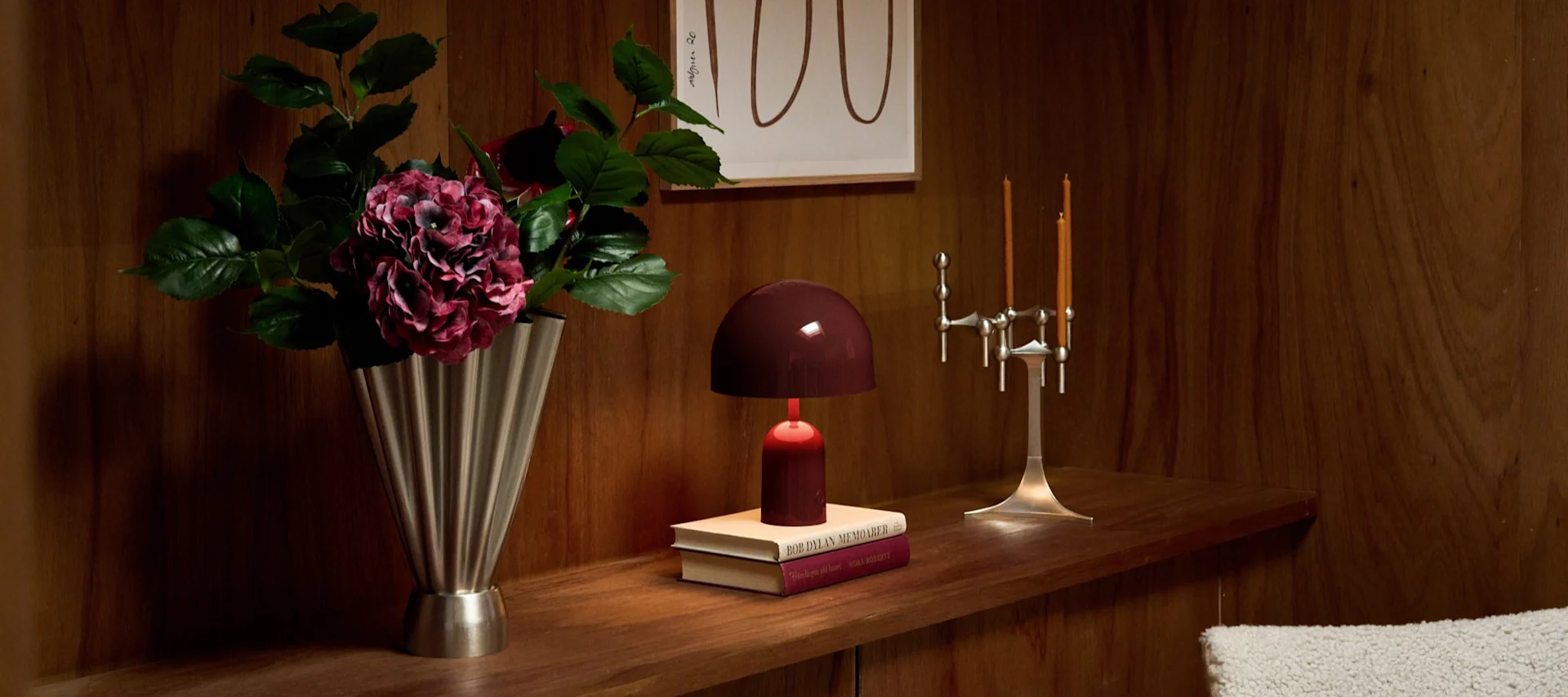 Holzregal: silberne Vase mit dunklen Hortensien, burgunderfarbene Lampe auf Büchern, silberner Kerzenhalter mit orange Kerzen.