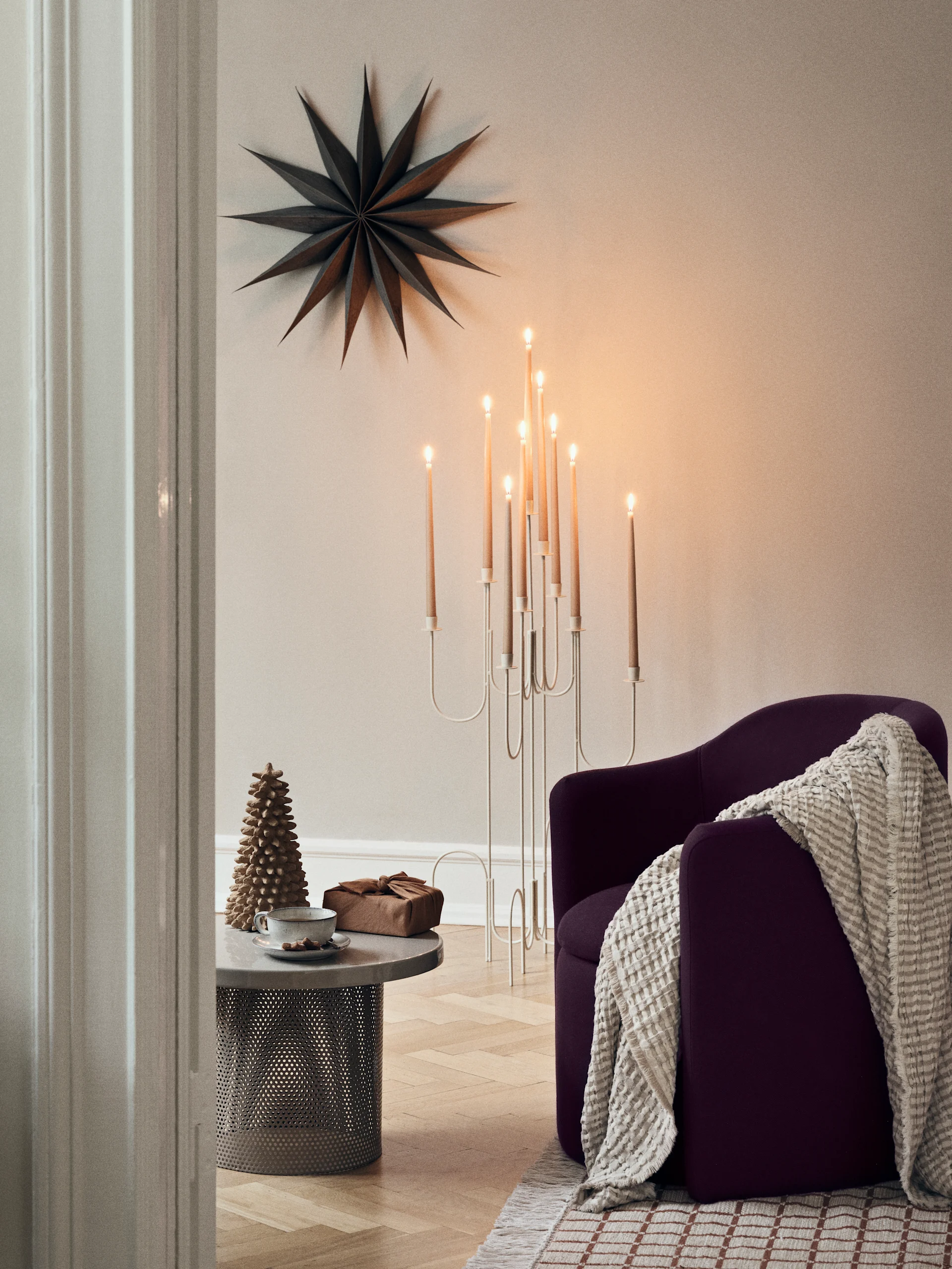 Eine helle Winterlandschaft durch ein Fenster, mit schneebedeckten Bäumen und einem Sonnenstrahl. Innen, ein Tisch mit Kerzen und Papierbäumen, und ein Papierstern hängt im Fenster.