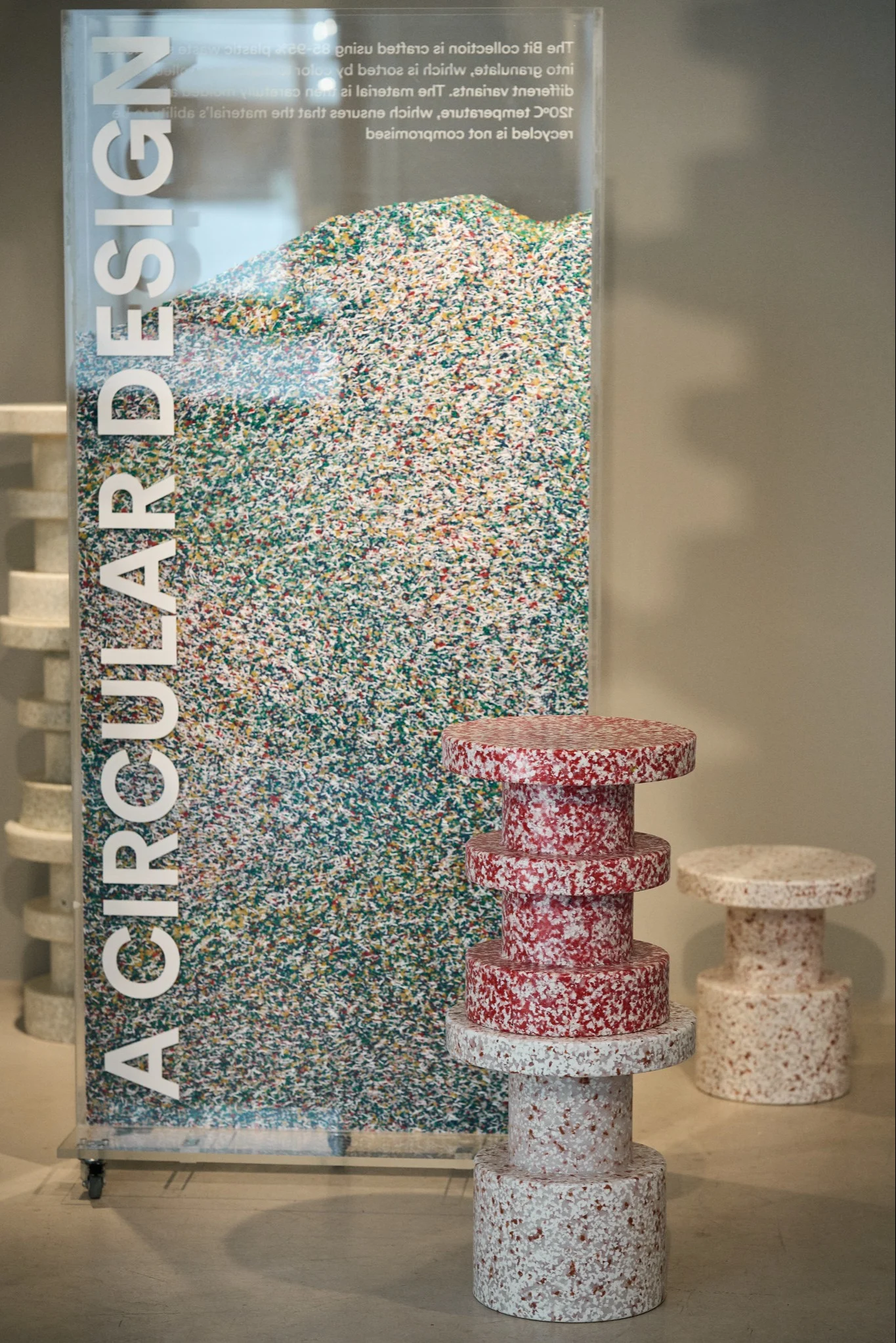 Ausstellung mit klarem Kasten aus bunten Plastikflocken und Text "A CIRCULAR DESIGN", daneben gesprenkelte rot-weiße und hellbeige Hocker aus recycelten Materialien.