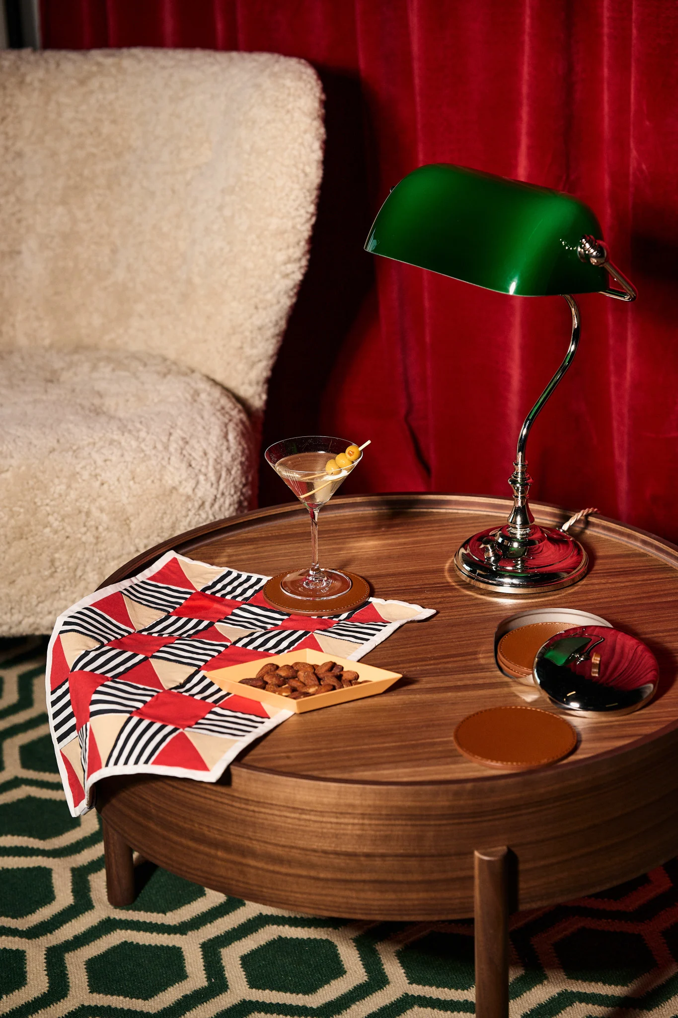 Martini, Mandeln, grüne Bankerlampe auf Holztisch mit geometrischem Tuch, cremefarbenem Sessel und roten Vorhängen.