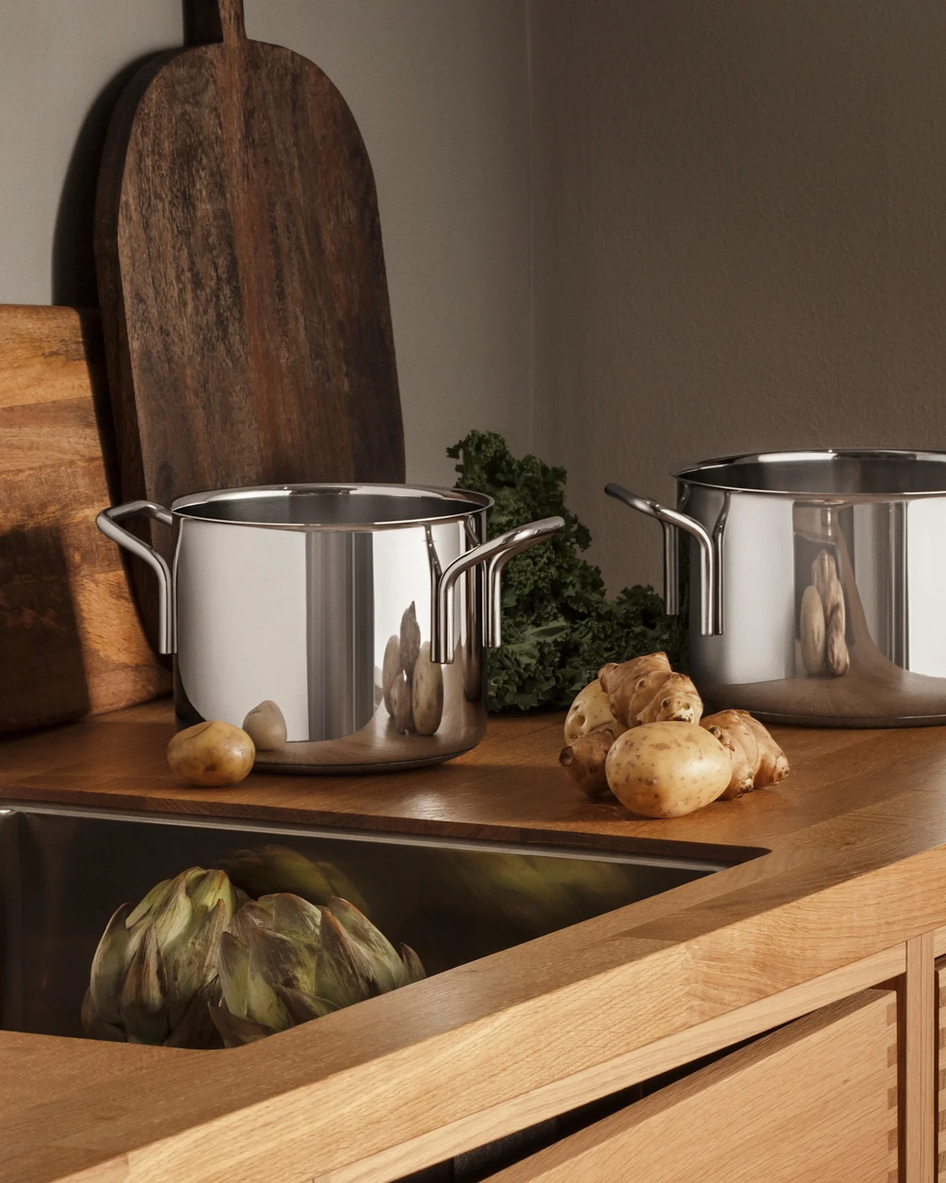 Moderne Küche mit Holztheke, Edelstahlspüle, Wasserhahn, drei Töpfen und frischen Produkten wie Kartoffeln und Artischocken.