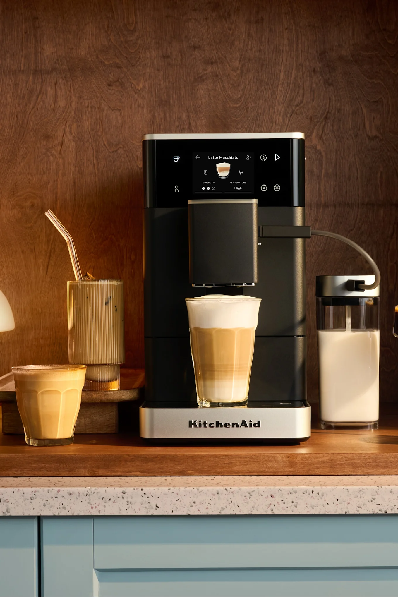 KitchenAid Espressomaschine füllt Latte Macchiato in hohes Glas, daneben andere Kaffees und Milch.