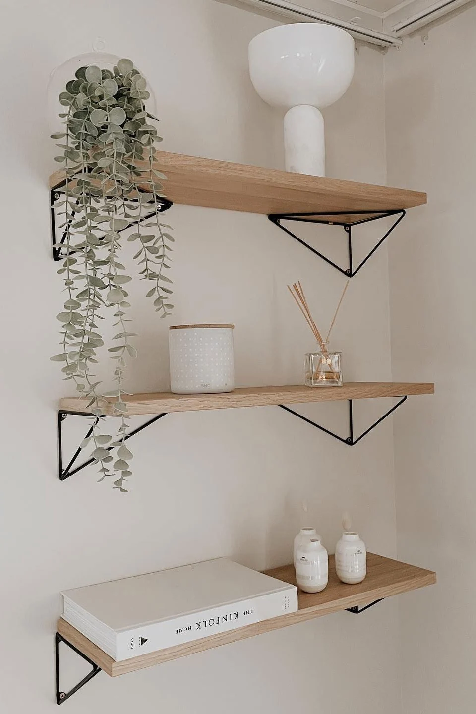 Drei helle Holz-Schwebregale mit schwarzen dreieckigen Halterungen. Zu den Gegenständen gehören eine Hängepflanze, eine weiße Vase, ein gepunkteter Behälter, ein Diffusor, ein weißes Buch und kleine weiße Vasen.