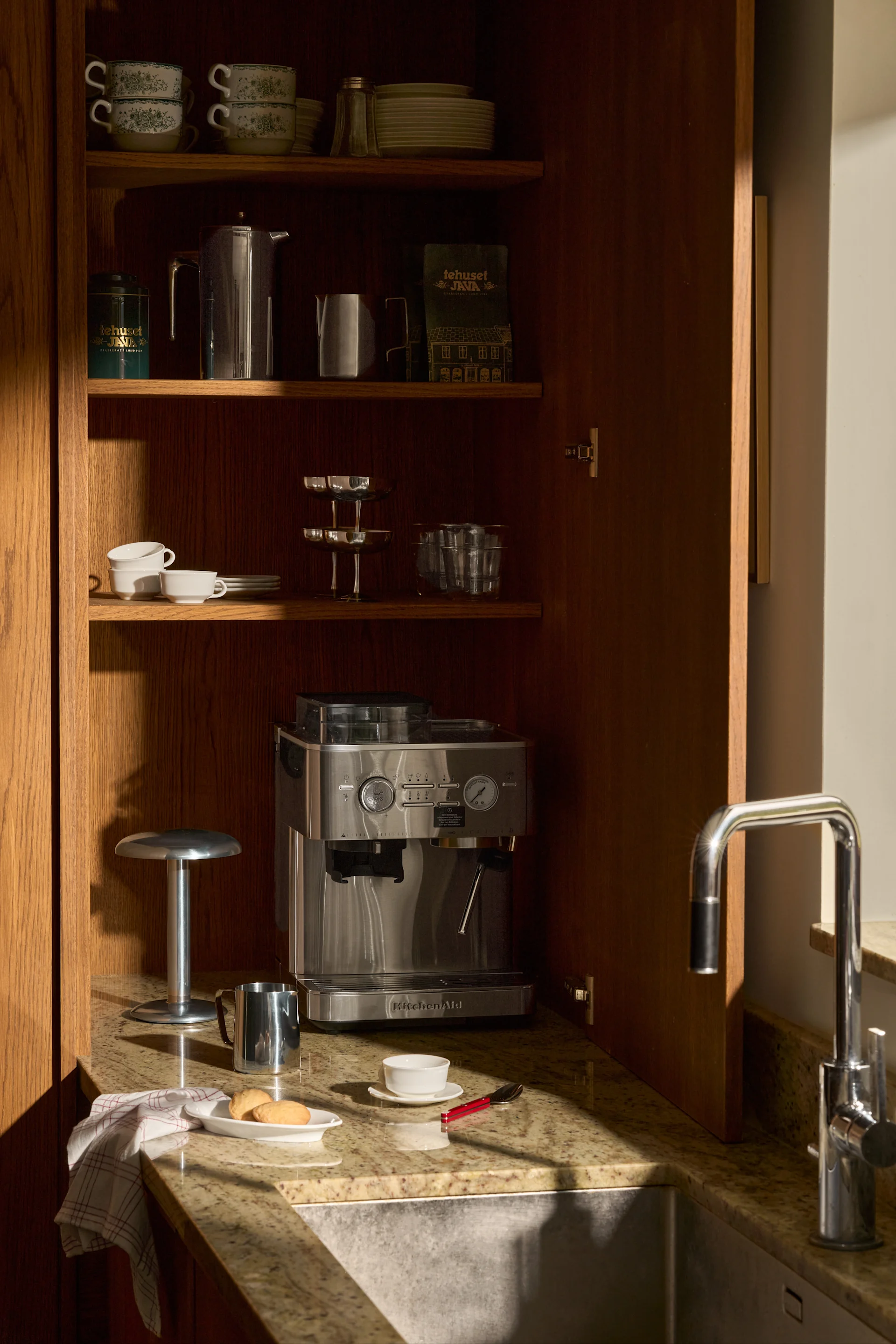 Offener Holzschrank mit KitchenAid Espressomaschine, gestapelten Tassen, Tellern, Kaffeeutensilien und Spüle auf Granitplatte.