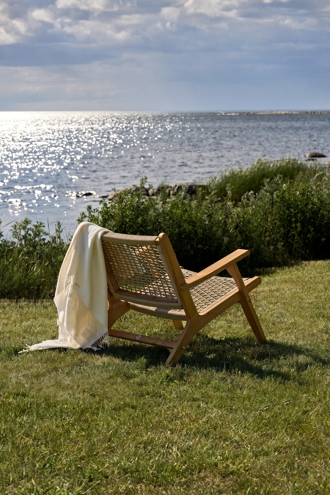 Hölzerner Loungesessel mit einer weißen Decke auf grünem Gras, mit Blick auf glitzerndes Wasser unter einem bewölkten Himmel.