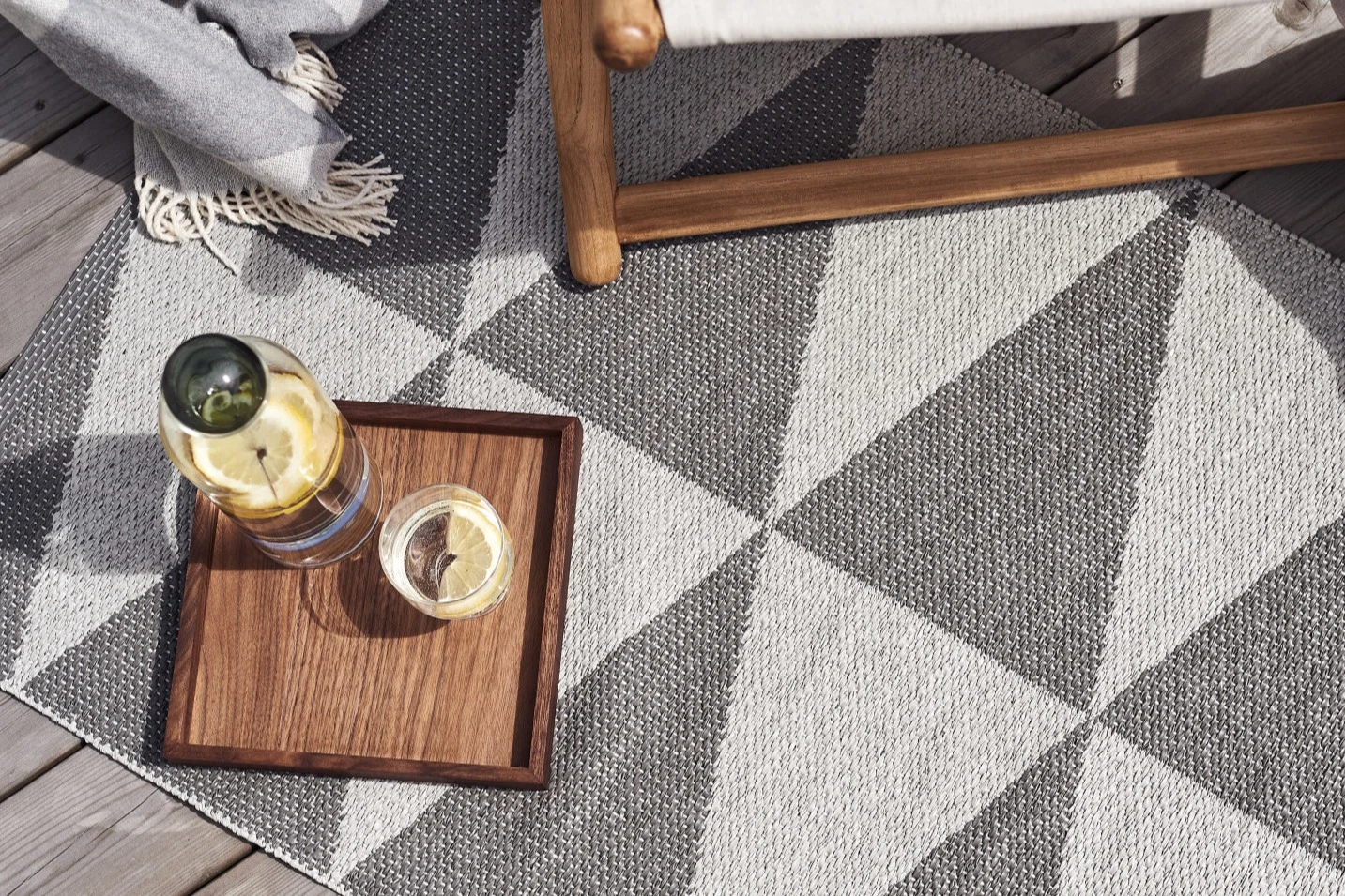 Geometrischer grau-weißer Outdoor-Teppich mit einem Holztablett mit Zitronenwasser, neben einem Stuhl auf einer Terrasse.