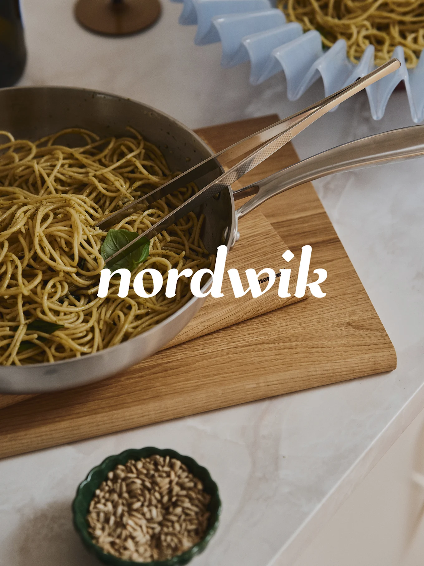 Spaghetti mit Pesto in einer Stahlpfanne mit Zange auf einem Holzbrett. Eine Schale mit Samen ist unten, ein Nudelgestell im Hintergrund. 'nordwik' Textüberlagerung.